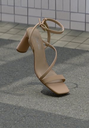 Beige høyhælte sandal med remmer og blokkhæl som svever over asfalt ved flislagt veggbakgrunn.