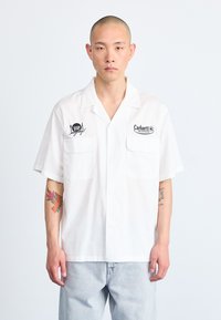 Camisa blanca de manga corta hecha de algodón. Presenta dos bolsillos frontales y logotipos bordados en el lado izquierdo del pecho. Jeans de mezclilla azul claro.
