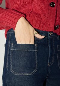 Červený pletený sveter s rebrovanou textúrou a veľkými gombíkmi, skombinovaný s tmavomodrými džínsami, ktoré majú viditeľné švy a ruku v vrecku.