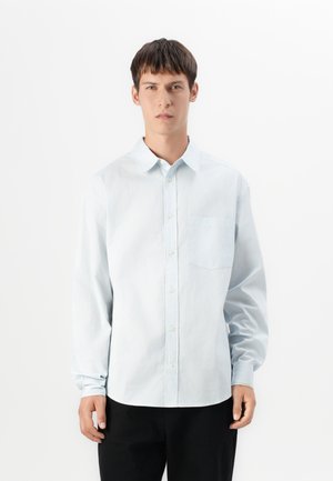Jonge man met kort donker haar draagt een lichtblauw overhemd met knopen en zwarte broek, staand tegen een effen witte achtergrond.