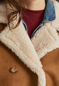 Brun mockasinsjacka med en krämfärgad fuskpälskrage, denimdetaljer och knappstängning. Har en mjuk textur och ett lagerpå-lager kragdesign.