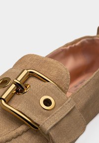 Gros plan sur un loafer beige texturé avec une grande boucle dorée brillante et des œillets sur la bride avant de la chaussure.