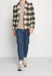 Schwarz-weiß kariertes Sakko über einem beigen Pullover, kombiniert mit blauen Jeans und beigen Sneakers. Mit klassischem Schnitt und umgeschlagenen Ärmeln.