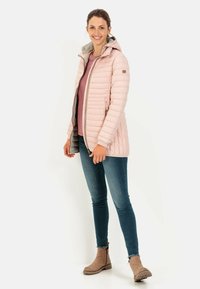 Veste rose rembourrée avec une capuche, fermeture éclair et doublure grise, portée sur un haut violet et un jean bleu, associée à des bottes de cheville beiges.