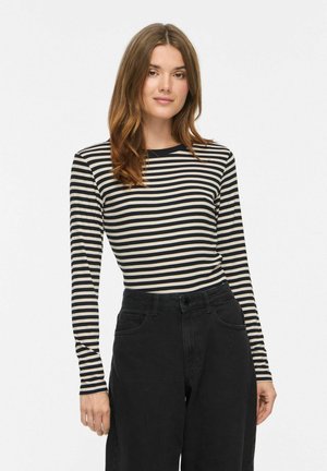 Donna con capelli castani fino alle spalle che indossa una maglietta a righe bianche e nere a maniche lunghe e pantaloni neri a vita alta su sfondo bianco.