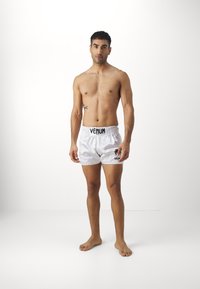 White satin shorts with elastic waistband, featuring black "Venum" branding. The model is barefoot and shirtless, posing frontally.