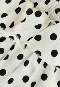 Vitt tyg med framträdande svarta prickar, med en strukturerad yta och volangkanter, som skapar en lagerpålagd, mönstrad design.