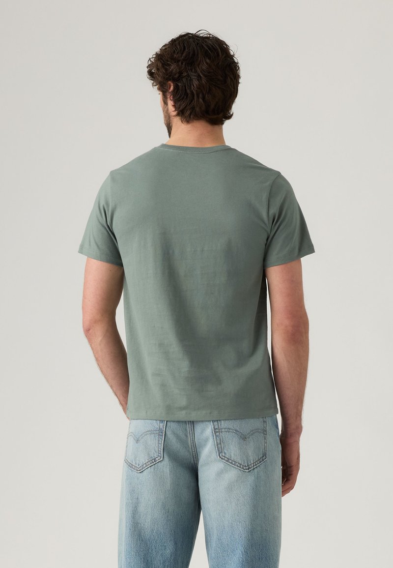 Man with dark curly hair wearing a muted green short-sleeve t-shirt and light blue denim jeans, facing away against a plain background.