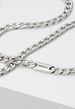 Hopeanvärinen ketjukaulakoru, jossa on litteitä toisiinsa lukittuvia lenkkejä ja suorakaiteen muotoinen kaiverrettu merkki, jossa lukee "BREIL", valkoisella taustalla.