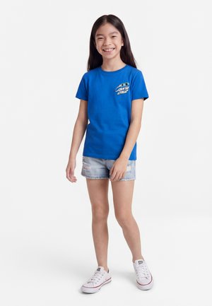 Jong meisje lacht, draagt een blauw Santa Cruz T-shirt, licht gerafelde spijkershorts en witte sneakers, staand tegen een witte achtergrond.