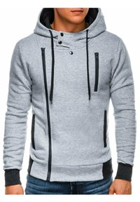 Sweat à capuche gris avec fermeture éclair à l'avant, accents noirs, et capuche à cordon de serrage. Comprend des poches de chaque côté ainsi que des poignets et un ourlet côtelés.