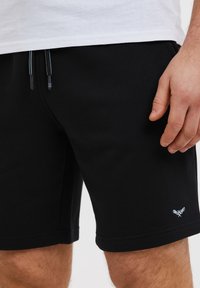 Shorts noirs en mélange de coton, dotés d'une taille à cordon de serrage et d'un petit logo bleu clair sur la jambe inférieure. Texture douce, longueur mi-cuisse.