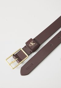 Calvin Klein odinė diržas tamsiai burgundiškai su auksiniu kvadratiniu sagtimi. Jame yra mažas įspaustas logotipas ant diržo kilpos ir tvarkingos briaunos.
