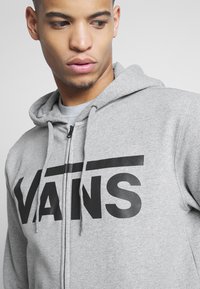 Szara bluza z kapturem wykonana z polaru, z czarnym logo "VANS". Zawiera zamek błyskawiczny z przodu oraz akcenty w postaci sznurka na kapturze.