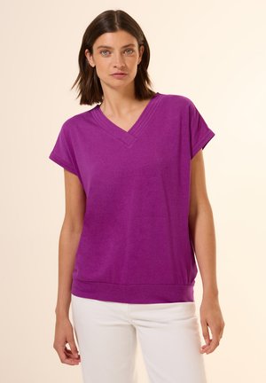 T-shirt violet à col en V, à manches courtes, en tissu doux, coupe décontractée, col côtelé, porté avec un pantalon blanc, texture lisse, sans motifs.