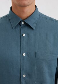 Teal overhemd met knopen van lichtgewicht stof, met een puntkraag, een borstzak en contrasterende witte knopen op een glad oppervlak.