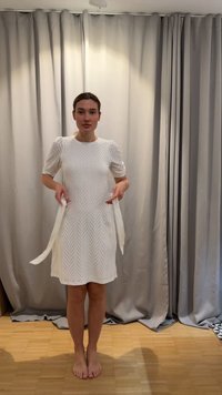 Sieviete stāv basām kājām uz koka grīdas, valkājot baltu ceļgalu garu kleitu ar pūkainām piedurknēm, rokās turot divas auduma lentes.