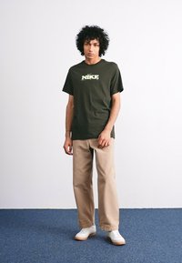 Mørkegrøn bomulds T-shirt med gult Nike-logo, parret med beige brede bukser og hvide sneakers. Enkel, afslappet design.