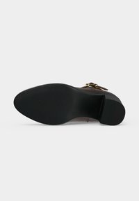 Semelle en caoutchouc noire avec avant et talon texturés, attachée à une chaussure en cuir marron avec un détail de boucle dorée.