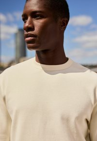 Lys beige tekstureret bluse med lange ærmer og rund halsudskæring. Stofmet har et subtilt rutemønster.