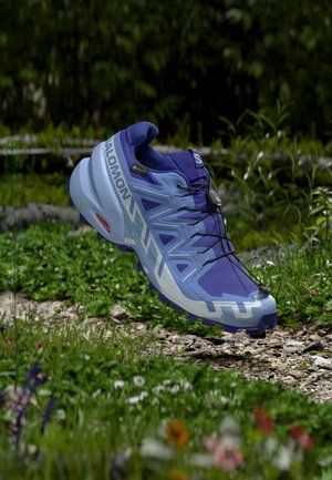 Scarpa da trail running Salomon blu e viola con Gore-Tex e sistema di allacciatura rapida che fluttua sopra un sentiero di ghiaia circondato da piante verdi e fiori.