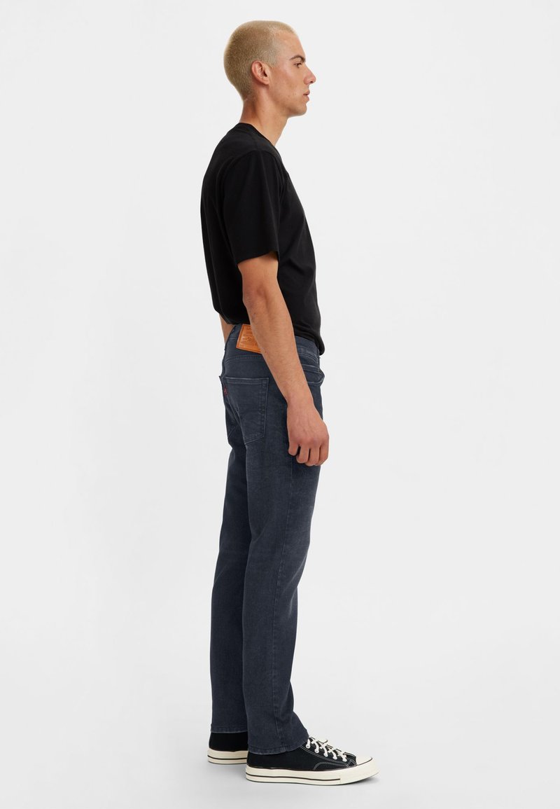 Jean bleu foncé en denim, avec une coupe slim, droite, et une patch en cuir marron à la ceinture arrière. Associé à un T-shirt noir et des baskets.