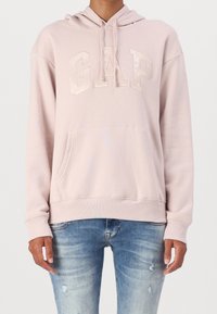 Sudadera con capucha de color rosa claro, con un bolsillo canguro frontal, que presenta un logo de GAP texturizado. Tejido suave con un ajuste relajado y dobladillo acanalado.