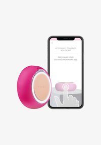Dispositif facial en silicone rose avec un design circulaire et un logo sur le devant, affiché aux côtés d'un smartphone montrant les instructions d'installation.