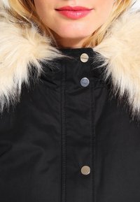 Svart vinterjacka med huva med pälsdetaljer, silvriga tryckknappar, texturerat tyg och vertikala sömdetaljer.