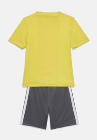 Camiseta deportiva amarilla de manga corta y cuello redondo, combinada con pantalones cortos grises que tienen rayas blancas a los lados y una cinturilla elástica.
