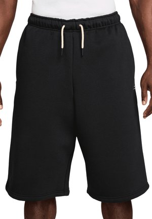 Schwarze, knielange Sportschuhe mit weißem Kordelzugbund, getragen von einer stehenden Person mit dunkler Hautfarbe und weißem Hemd.