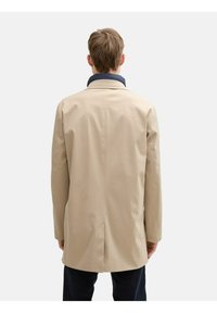 Beige regenjas met een marineblauwe kraag, heeft een rechte snit, lange mouwen en een split aan de achterkant voor extra bewegingsvrijheid.