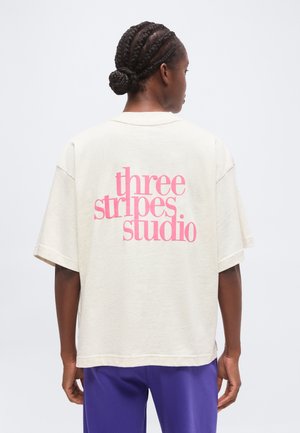 Person med flettet hår i en knold iført en løs off-white t-shirt med pink "three stripes studio" tekst på bagsiden og lilla bukser.