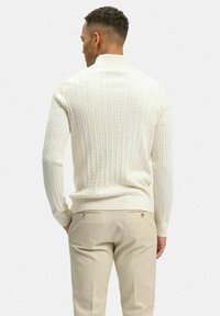 Vit kabelstickad tröja med ribbade ärmslut och rund halsringning, kombinerad med ljusbeiga byxor med två blixtlåsfickor bak.