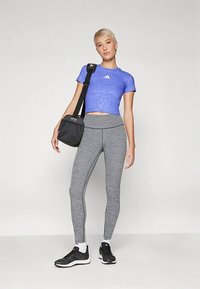 Egy személy kék Adidas crop topot, szürke leggingset, fekete sportcipőt visel, és egy fekete Adidas válltáskát hordoz egy sima fehér háttér előtt.