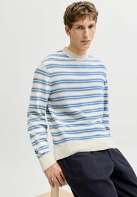 Jeune homme assis sur un tabouret, portant un pull rayé crème et bleu ainsi que un pantalon foncé, sur un fond clair uni.