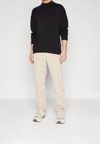 Zwarte coltrui, crèmekleurige corduroy broek en beige sneakers. De trui heeft lange mouwen en een relaxed fit; de broek heeft een ribbelstructuur.