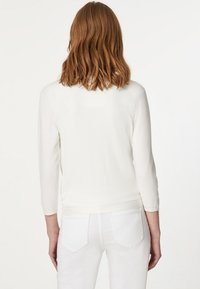 Suéter blanco de punto con mangas tres cuartos, textura suave y dobladillo redondeado, combinado con jeans blancos, mostrado desde la parte de atrás.