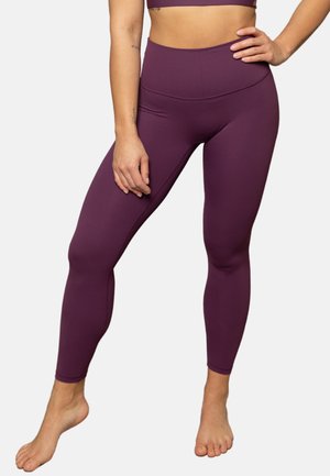 Woman wearing high-waisted, purple leggings and matching sports bra, standing barefoot with one hand on hip against a plain background.