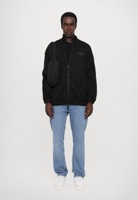 Giacca nera con zip e colletto alto, polsini aderenti e un piccolo patch con logo, indossata sopra una maglietta nera e abbinata a jeans azzurri chiari e sneakers bianche.