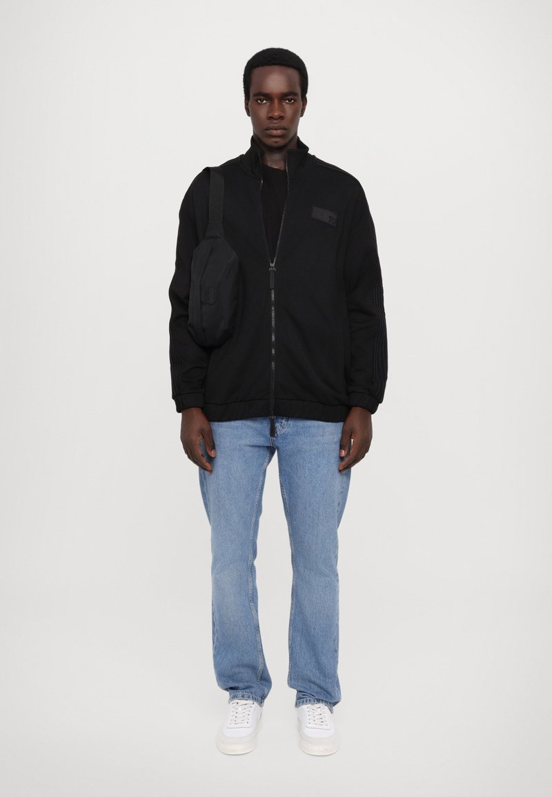 Giacca nera con zip e colletto alto, polsini aderenti e un piccolo patch con logo, indossata sopra una maglietta nera e abbinata a jeans azzurri chiari e sneakers bianche.