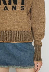 Pull en maille marron avec des accents côtelés et un logo noir, associé à une jupe en jean bleue avec un ourlet à franges.