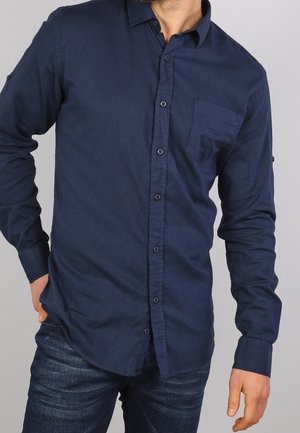 Homme portant une chemise bleu marine à boutons avec poche poitrine et un jean bleu foncé, debout avec une main dans la poche.