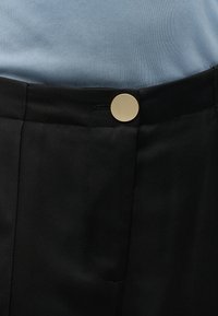 Κοντινό πλάνο στη μέση μαύρου παντελονιού με μεγάλο στρογγυλό χρυσό κουμπί και μέρος ενός ανοιχτόχρωμου μπλε πουκαμίσου που έχει μπει μέσα.