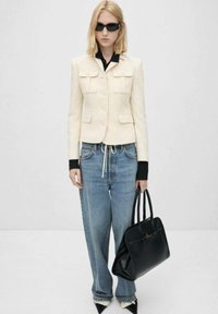 Chaqueta de tweed crema con bolsillos, combinada con jeans azules sueltos y un bolso negro. Camisa negra debajo y zapatos negros elegantes.