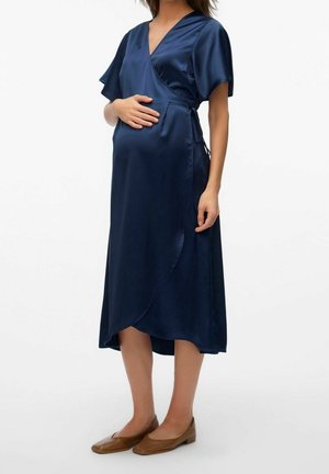 Femme enceinte portant une robe portefeuille bleu marine et des ballerines marron, une main reposant sur son ventre sur un fond uni.