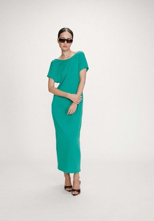 Femme portant une robe midi turquoise à épaules dénudées, des talons noirs ouverts à l\'avant et des lunettes de soleil foncées, debout les bras croisés devant un fond blanc.