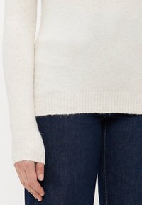 Personne portant un pull en tricot crème avec des poignets côtelés et un jean en denim bleu foncé, main détendue le long du corps.