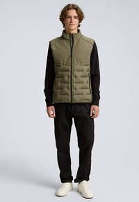 Ovitka olivno zelene barve z visokim ovratnikom, z zadrgo spredaj in črnimi dolgimi rokavi pod njo. Nošena s črnimi hlačami in belimi supergami.