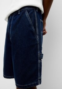 Pantalones cortos de mezclilla azul oscuro con costuras blancas, que cuentan con bolsillos laterales y un bolsillo tipo cargador en el muslo. Ajuste casual, longitud a mitad del muslo.